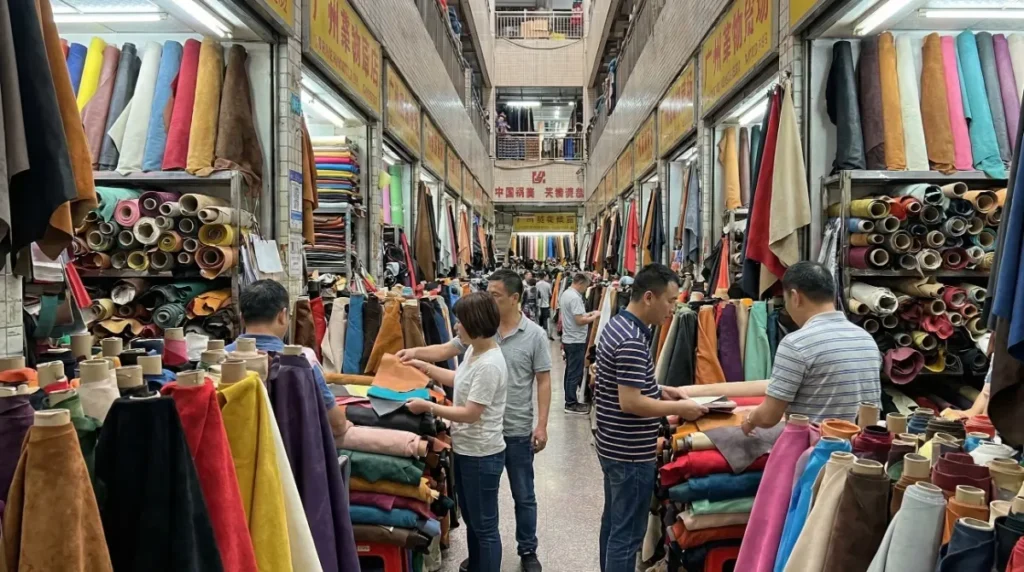 A crowded, vibrant outdoor wholesale market street in Guangzhou overflowing with stalls selling huge rolls of leather and textiles to buyers.