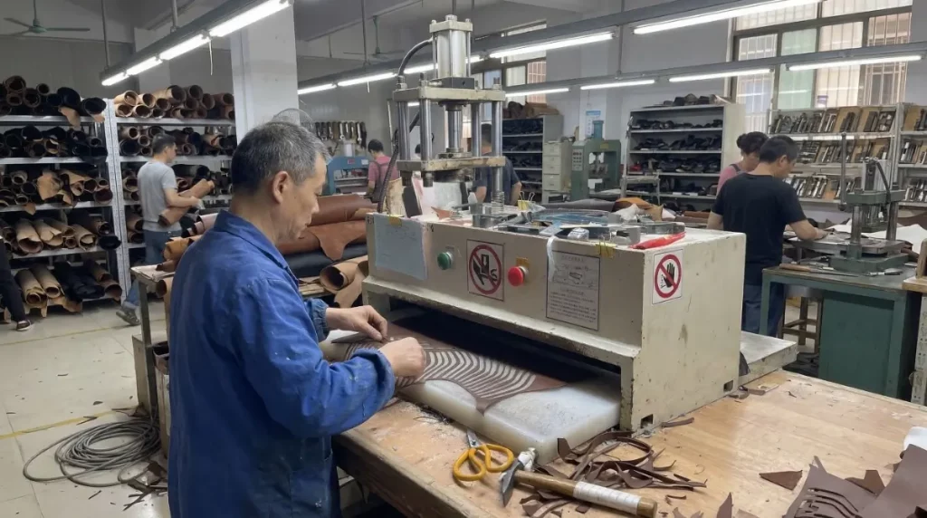 A factory worker in a blue coat meticulously feeding a piece of patterned leather into a large industrial hydraulic clicking press in a busy workshop.